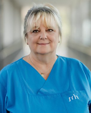 Eine Frau mit blonden Haaren trägt ein hellblaues medizinisches Outfit. Sie steht in einem hellen, modernen Raum und schaut direkt in die Kamera.