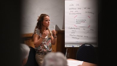 Eine Frau steht vor einem Flipchart und erklärt etwas, während Teilnehmer am Tisch zuhören. Notizen sind auf dem Flipchart sichtbar.