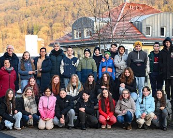 Eine Gruppe von etwa 40 Personen steht im Freien, umgeben von Bäumen und Gebäuden, und posiert für ein Gruppenfoto.
