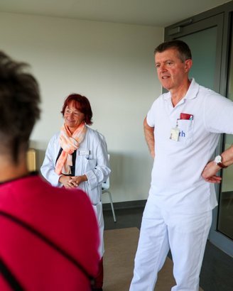 Zwei medizinische Fachkräfte stehen in einem Raum. Eine Frau mit rotem Haar trägt einen weißen Kittel und einen Schal, der Mann in weißer Kleidung hat seine Hände in die Hüften gestützt.