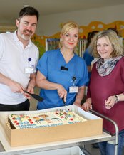 Drei Personen schneiden gemeinsam eine große Torte auf einem Wagen an, während sie in einem hellen Raum stehen.