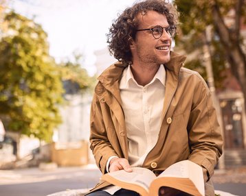 Ein junger Mann mit lockigem Haar und Brille sitzt im Freien, trägt eine beige Jacke und ein weißes Hemd. Er hält ein aufgeschlagenes Buch und lächelt, während er in die Ferne blickt.