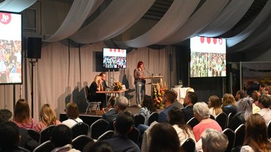 Eine Rednerin steht an einem Podium vor einem Publikum, während auf Bildschirmen im Hintergrund eine Veranstaltung gezeigt wird.