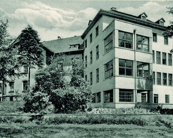 Eine historische Schwarz-Weiß-Postkarte zeigt die Gartenansicht eines mehrstöckigen Gebäudes mit großen Fenstern, umgeben von Bäumen und Sträuchern. Im Hintergrund ist ein weiteres Gebäude sichtbar.