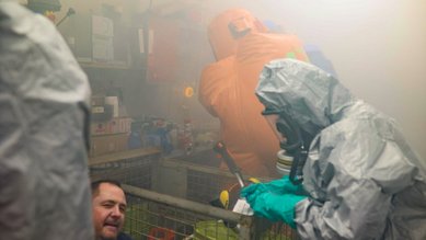 Ein Rettungsteam in Schutzanzügen hilft einer Person in einem verrauchten Raum, während Ausrüstung im Hintergrund sichtbar ist.