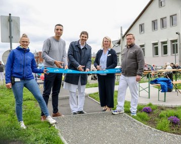 Eröffnung eines neuen Bereichs mit fünf Personen, die ein blaues Band durchschneiden. Im Hintergrund sind Tische und Stühle aufgestellt.