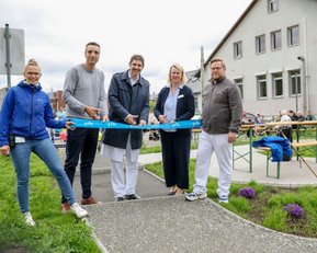 Eröffnung eines neuen Bereichs mit fünf Personen, die ein blaues Band durchschneiden. Im Hintergrund sind Tische und Stühle aufgestellt.