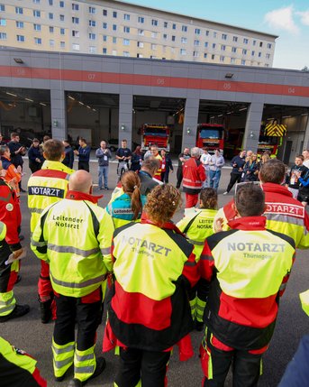 Eine Gruppe von Rettungskräften in leuchtenden Uniformen versammelt sich vor einem Gebäude, während sie einer Präsentation oder Besprechung zuhören. Im Hintergrund sind Feuerwehrfahrzeuge und andere Personen zu sehen.