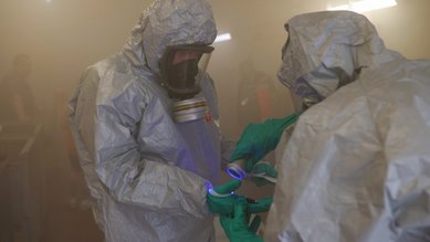 Zwei Personen in Schutzanzügen arbeiten zusammen, während sie ein fluoreszierendes Objekt in einer nebligen Umgebung untersuchen.