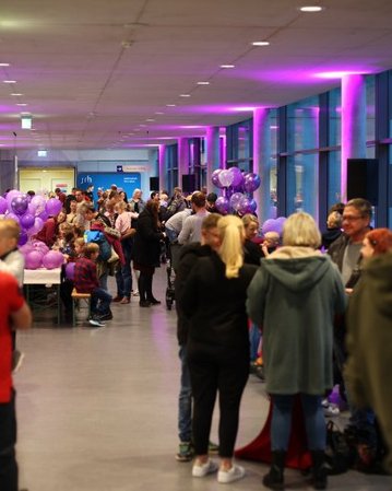 Eine Menschenmenge versammelt sich in einem langen Flur, geschmückt mit lila Ballons und Beleuchtung, während Tische mit Snacks und Getränken bereitstehen.