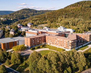 Ein Luftbild eines modernen Gebäudekomplexes, umgeben von Bäumen und Hügeln. Die Architektur besteht aus mehreren roten Ziegelgebäuden, die in einer U-Form angeordnet sind, mit Grünflächen und Straßen in der Nähe.