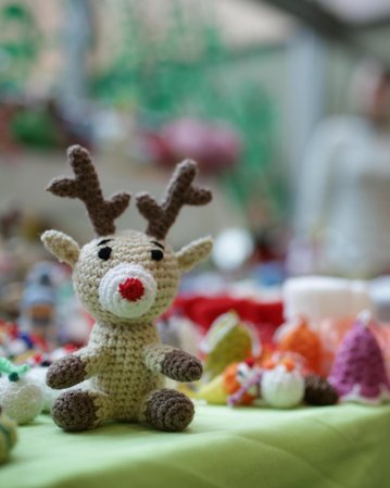 Häkelrentier mit roten Nasen und Geweih steht auf einem Tisch, umgeben von bunten Handwerksartikeln. Im Hintergrund sind zwei Personen unscharf zu sehen.