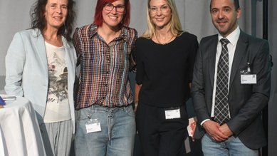 Vier Personen stehen zusammen, lächelnd, in einem Veranstaltungsraum. Sie tragen formelle Kleidung und haben Namensschilder an.