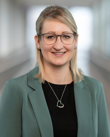 Eine Frau mit blonden Haaren trägt eine grüne Blazerjacke und eine schwarze Bluse. Sie lächelt und steht in einem hellen, modernen Raum.