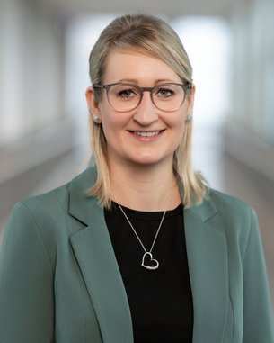 Eine Frau mit blonden Haaren trägt eine grüne Blazerjacke und eine schwarze Bluse. Sie lächelt und steht in einem hellen, modernen Raum.