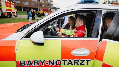 Ein Rettungswagen mit der Aufschrift "BABY-NOTARZT". Zwei Personen sitzen im Fahrzeug, während eine dritte Person außen steht.