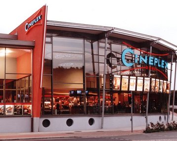 Cineplex-Gebäude mit moderner Architektur, großen Fenstern und auffälligem Logo. Plakate und Anzeigen sind sichtbar, die auf Filme hinweisen. Der Eingangsbereich ist gut beleuchtet und einladend.