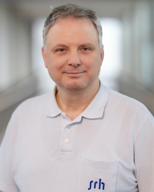 Mann mit kurzen grauen Haaren trägt ein helles Polohemd mit dem Logo "srh". Er steht in einem modernen Innenraum.