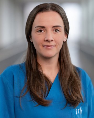 Frau mit langen, braunen Haaren trägt ein blaues Oberteil. Der Hintergrund ist unscharf und zeigt einen modernen Innenraum.