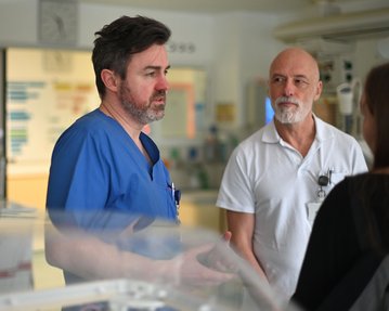 Zwei Ärzte im Gespräch, einer in blauer Arbeitskleidung und der andere in weißem Hemd, während eine Person ihnen zuhört.