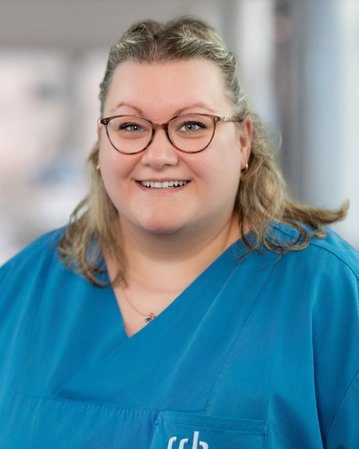 Eine lächelnde Frau mit Brille trägt ein blaues Oberteil mit dem Schriftzug "srh". Der Hintergrund ist unscharf und hell.