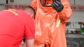 Ein Feuerwehrmann in einem orangefarbenen Schutzanzug wird von einem Kollegen bei der Ankleidung unterstützt.