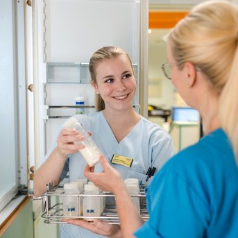 Eine Pflegekraft lächelt, während sie eine Flasche aus einem Kühlschrank hält. Eine andere Pflegekraft steht ihr gegenüber und schaut freundlich zu. Beide tragen medizinische Kleidung in hellblau und türkis.
