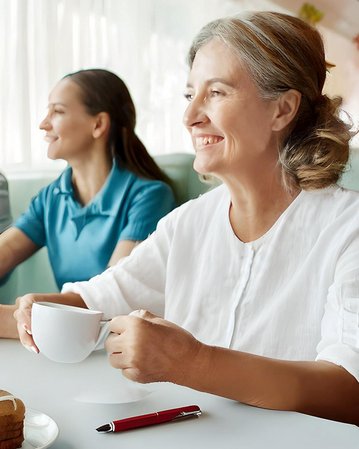 Eine ältere Frau mit grauen Haaren hält eine Tasse in der Hand und lächelt. Neben ihr sitzt eine jüngere Frau in einem blauen Shirt. Auf dem Tisch liegt ein Stück Kuchen und ein roter Stift.