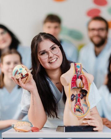 Eine lächelnde Frau hält anatomische Modelle in der Hand, während sie von mehreren Personen in medizinischer Kleidung umgeben ist.