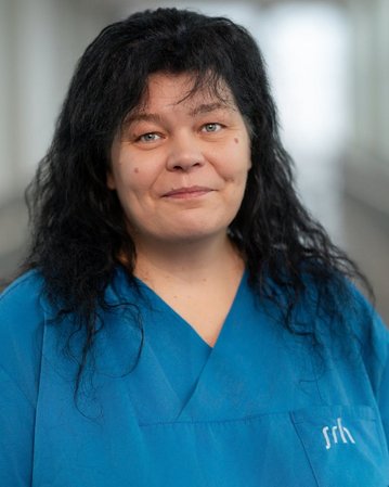 Frau mit langen, schwarzen Haaren trägt ein blaues Oberteil. Sie steht in einem hellen Flur und schaut direkt in die Kamera.