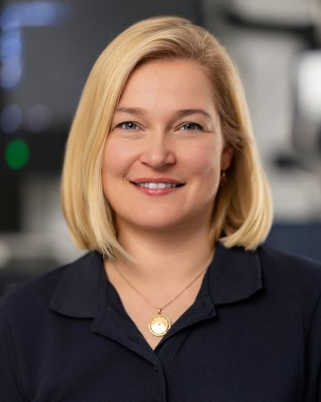 Eine blonde Frau mit kurzem Haar lächelt in die Kamera, trägt ein dunkles Oberteil und eine Halskette. Im Hintergrund sind medizinische Geräte sichtbar.