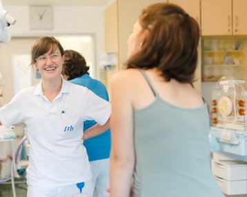 Eine Krankenschwester in einem Krankenhaus lächelt und spricht mit einer Patientin. Im Hintergrund sind medizinische Geräte und eine weitere Person zu sehen. Die Atmosphäre wirkt freundlich und professionell.