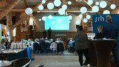 Eine Gruppe von Menschen sitzt in einem Veranstaltungsraum, während ein Referent auf einer Bühne eine Präsentation hält. Beleuchtung durch hängende Lampen.