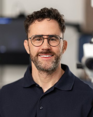 Ein Mann mit Brille und Bart lächelt in einem medizinischen Umfeld, während ein Mikroskop im Vordergrund sichtbar ist.