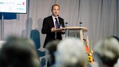 Ein Mann in Anzug spricht an einem Rednerpult während einer Veranstaltung, im Hintergrund sind Blumen und ein Bildschirm sichtbar.
