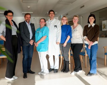 Eine Gruppe von sieben Personen in einem hellen Raum, darunter medizinisches Personal und Mitarbeiter, posiert freundlich für ein Foto.