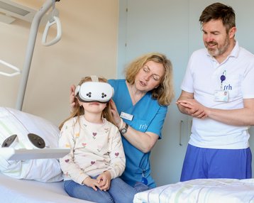 Ein Mädchen sitzt im Krankenhausbett und trägt eine VR-Brille, während eine Krankenschwester und ein Arzt ihr helfen.