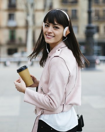 Junge Frau mit Kopfhörern und Kaffeebecher, die lächelnd in die Kamera schaut, während sie auf einem Platz steht.