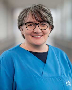 Eine lächelnde Frau mit kurzen grauen Haaren trägt einen blauen Kittel und eine Brille. Sie steht in einem hellen, modernen Raum.