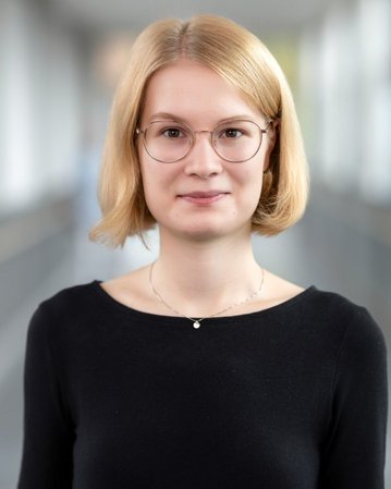 Junge Frau mit blonden, schulterlangen Haaren und Brille, trägt ein schwarzes Oberteil, steht in einem hellen Flur.