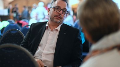Ein Mann im Anzug sitzt auf einem Stuhl und spricht mit einer Person, die ihm gegenüber sitzt. Der Hintergrund zeigt eine Veranstaltung mit mehreren Teilnehmern.