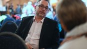 Ein Mann im Anzug sitzt auf einem Stuhl und spricht mit einer Person, die ihm gegenüber sitzt. Der Hintergrund zeigt eine Veranstaltung mit mehreren Teilnehmern.
