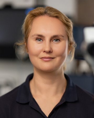 Eine Frau mit blonden Haaren lächelt in die Kamera, während sie vor einem medizinischen Gerät steht.