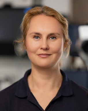 Eine Frau mit blonden Haaren lächelt in die Kamera, während sie vor einem medizinischen Gerät steht.