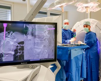 Ein Monitor zeigt bildgebende Verfahren während eines chirurgischen Eingriffs. Zwei Chirurgen in blauen OP-Kitteln und Masken arbeiten am Patienten, der sich hinter einem runden Gerät befindet.