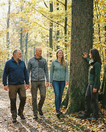 Vier Personen gehen auf einem Waldweg, während eine Frau an einem Baum steht und mit den anderen spricht. Herbstliche Umgebung mit buntem Laub.