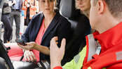 Drei Personen im Auto, eine Frau mit Mikrofon spricht, während zwei Frauen aufmerksam zuhören. Im Hintergrund sind weitere Personen sichtbar.