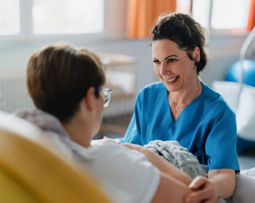 Eine Krankenschwester in blauer Uniform spricht lächelnd mit einer Patientin im Krankenhausbett.