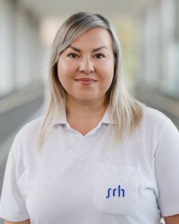 Frau mit langen, blonden Haaren trägt ein weißes Polohemd mit dem Logo "srh". Sie steht in einem hellen, modernen Innenraum.