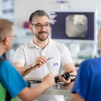 Ein Mann mit Brille und grauen Haaren erklärt etwas, während er ein medizinisches Gerät hält. Zwei Personen in blauen und grünen Oberteilen hören aufmerksam zu. Im Hintergrund ist ein Monitor sichtbar.